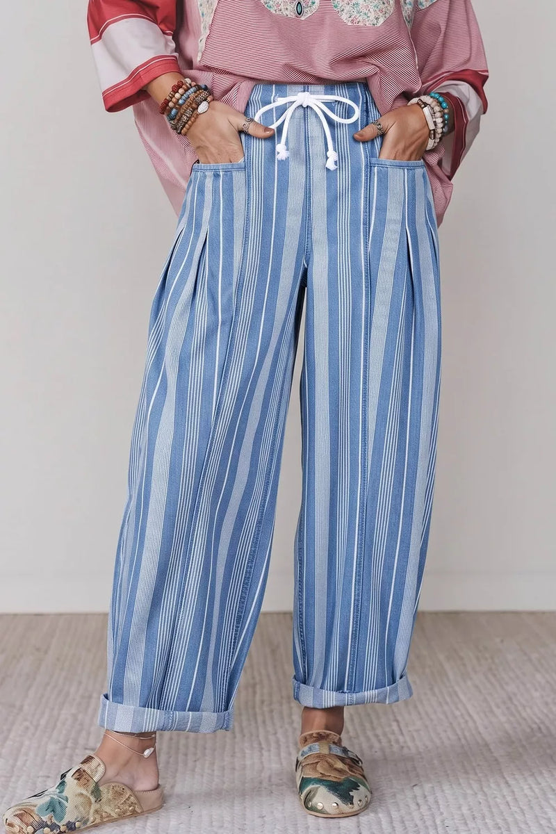 A person wearing loose blue and white vertically striped wide-leg pants with a white drawstring waist, paired with a pink patterned top and floral slip-on shoes, standing against a plain light wall.