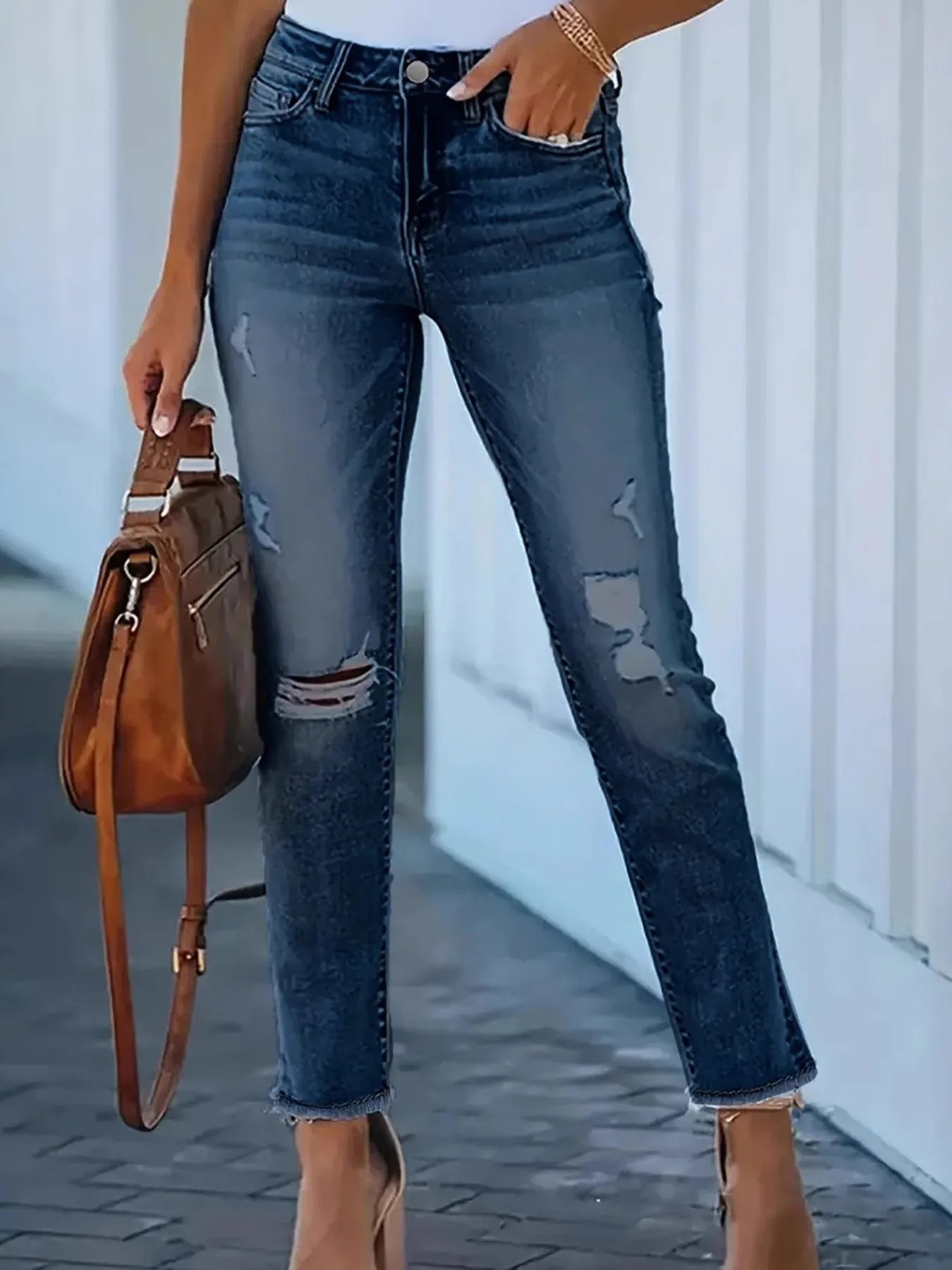 Person wearing distressed blue jeans holding a brown leather bag.