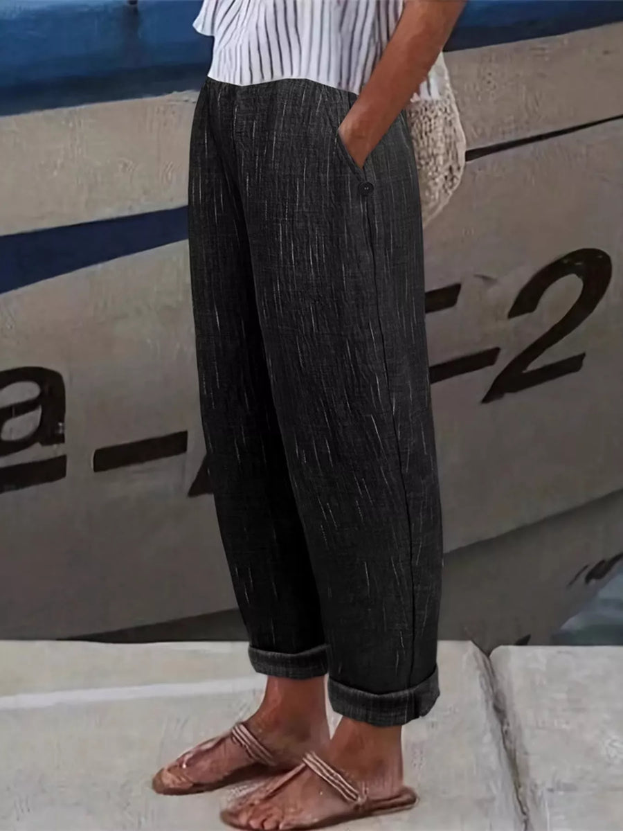 A person wearing dark charcoal gray textured wide-leg trousers with rolled cuffs, paired with a white striped top and flat sandals, standing in front of a boat.