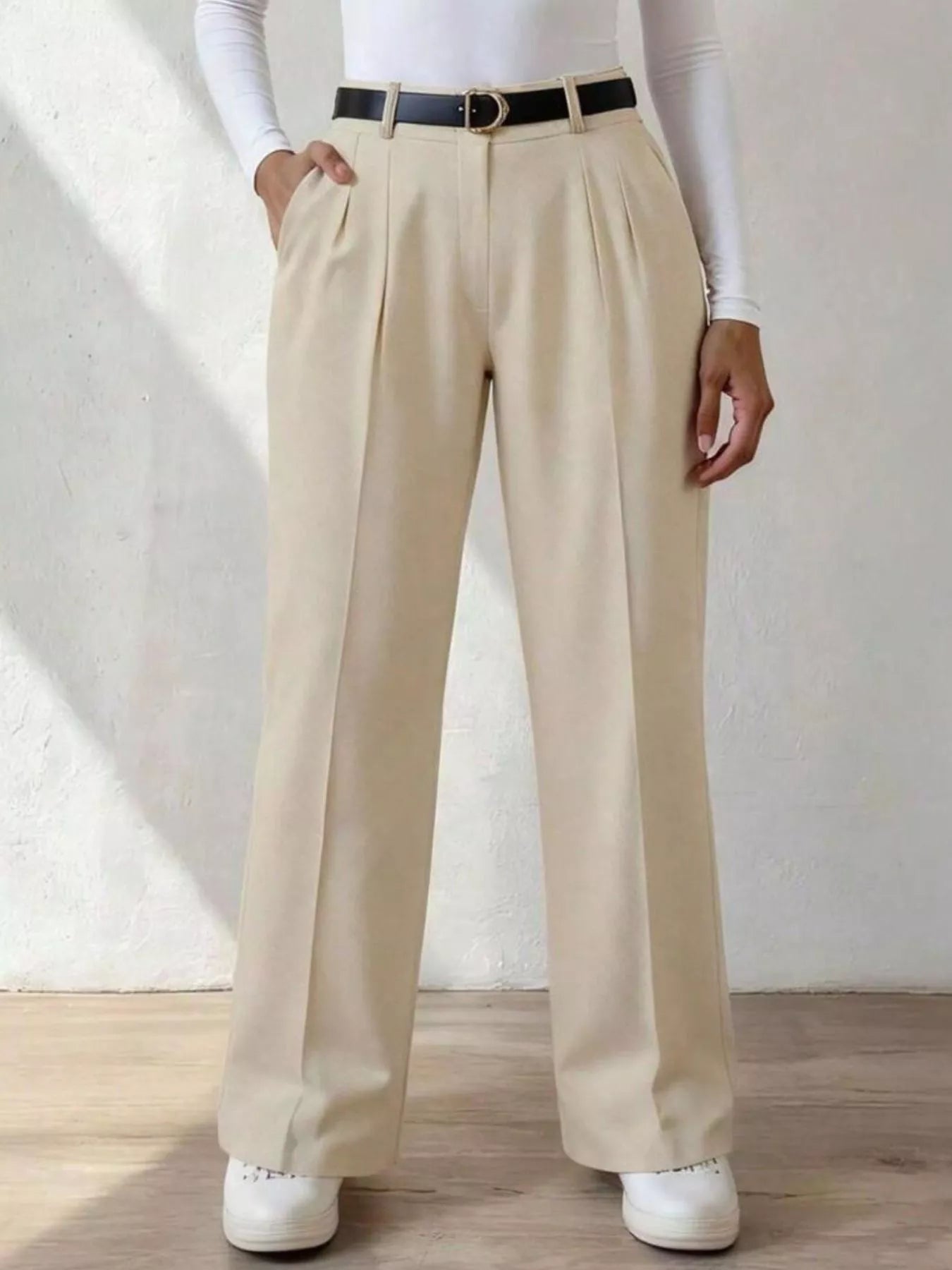 Person wearing beige pleated pants with a white shirt and white shoes on a light background