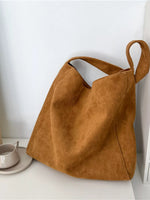 A large slouchy suede tote bag in a warm camel brown color, resting on a white surface next to a small ceramic cup and saucer against a white wall.