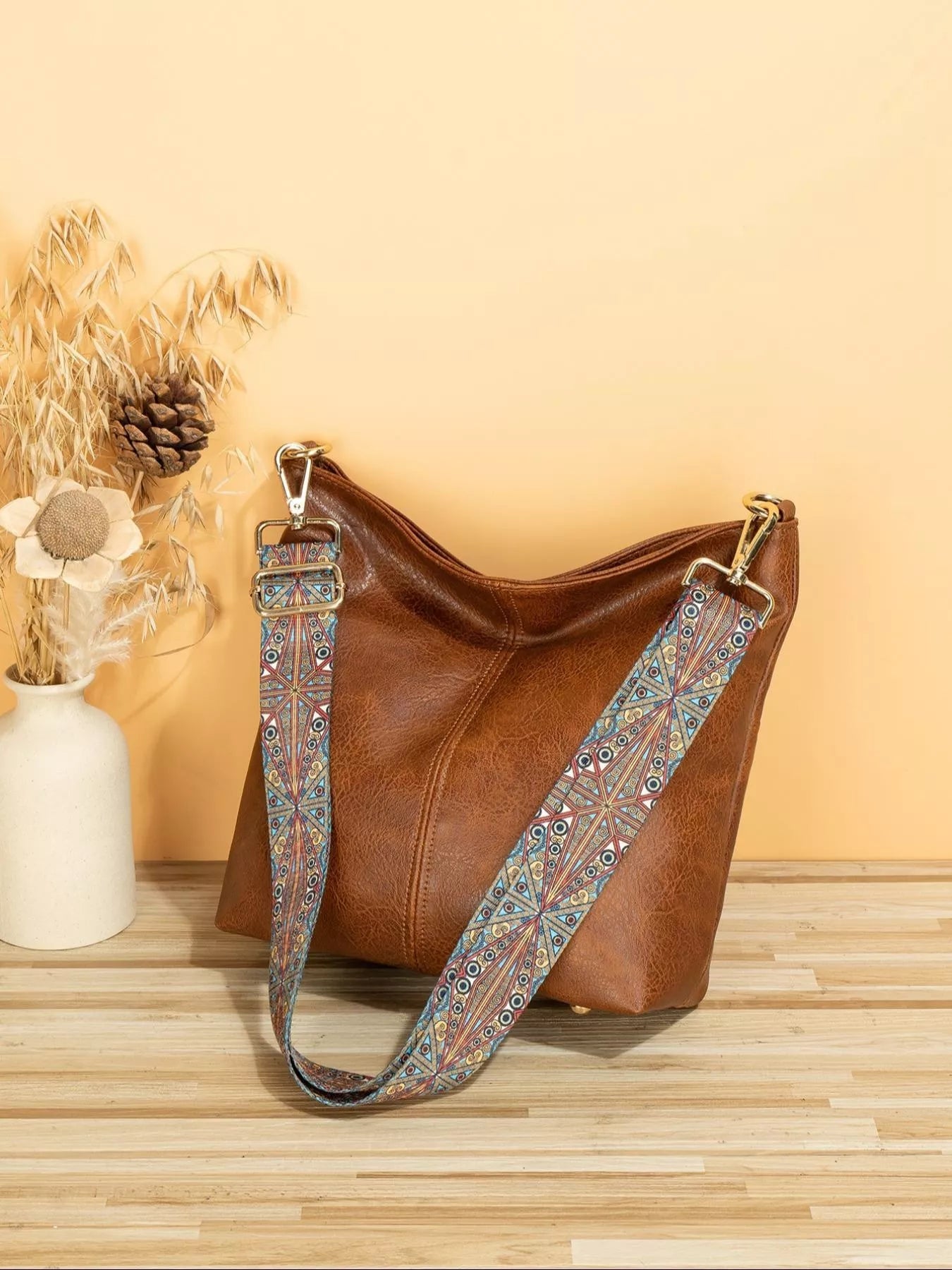 Brown leather handbag with a patterned strap on a wooden surface with a beige background