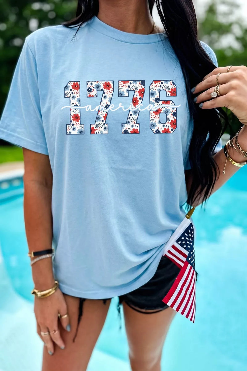 Person wearing a light blue t-shirt with '1776' design, holding an American flag, by a pool.