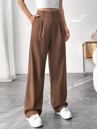 Person wearing brown wide-leg pants and white sneakers in a minimalistic room.