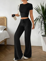 Person wearing a black outfit with wide-leg pants in a room with a bed and plant.