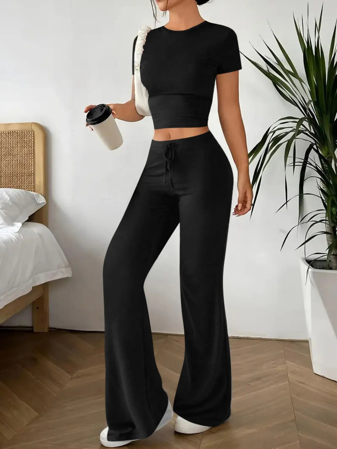 Person wearing a black outfit with wide-leg pants in a room with a bed and plant.