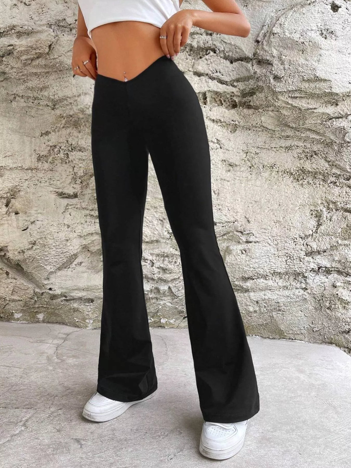 A person wearing black high-waisted flare pants with white sneakers, standing against a textured concrete wall.