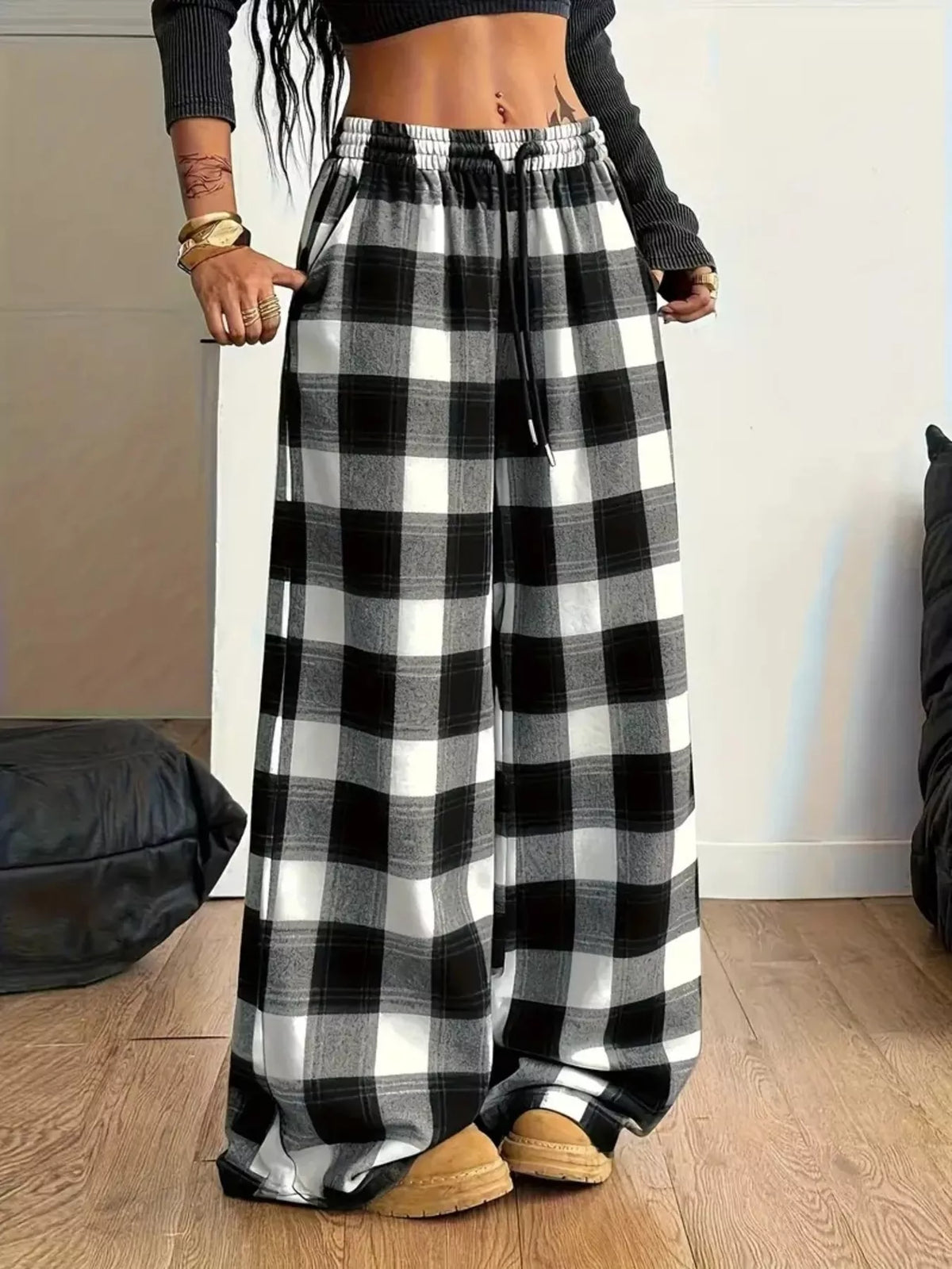 A person wearing black and white buffalo plaid wide-leg pants with an elastic drawstring waist, paired with a black long-sleeve crop top and tan slip-on shoes, standing on a wooden floor.