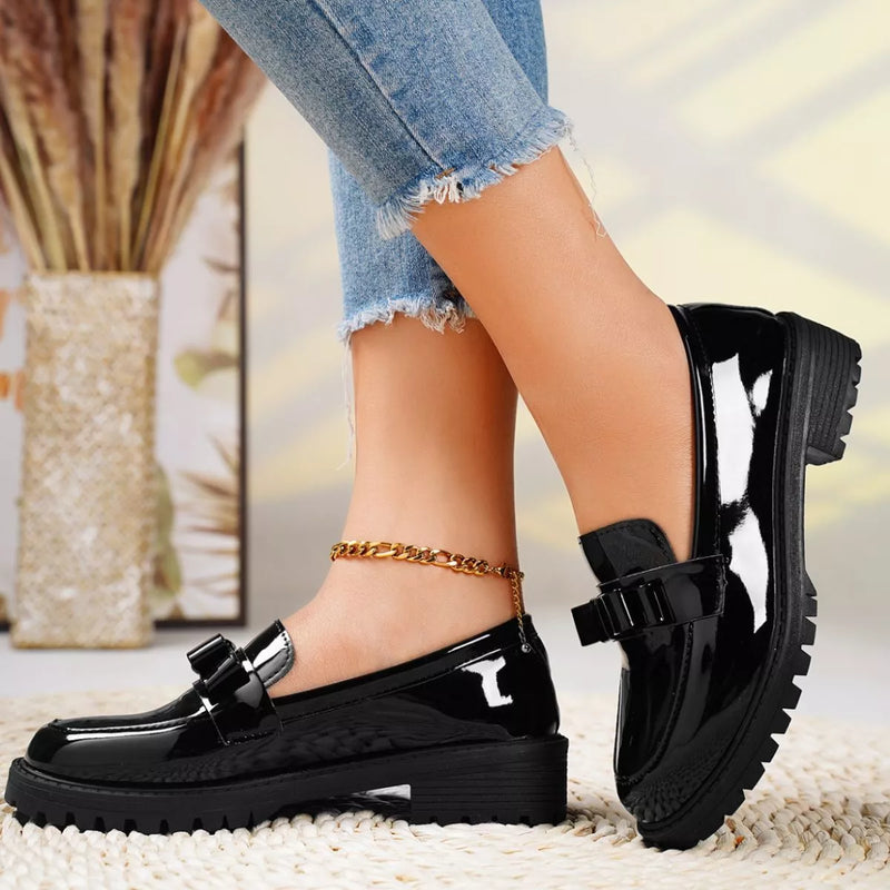Black patent leather shoes worn with denim jeans on a blurred indoor background