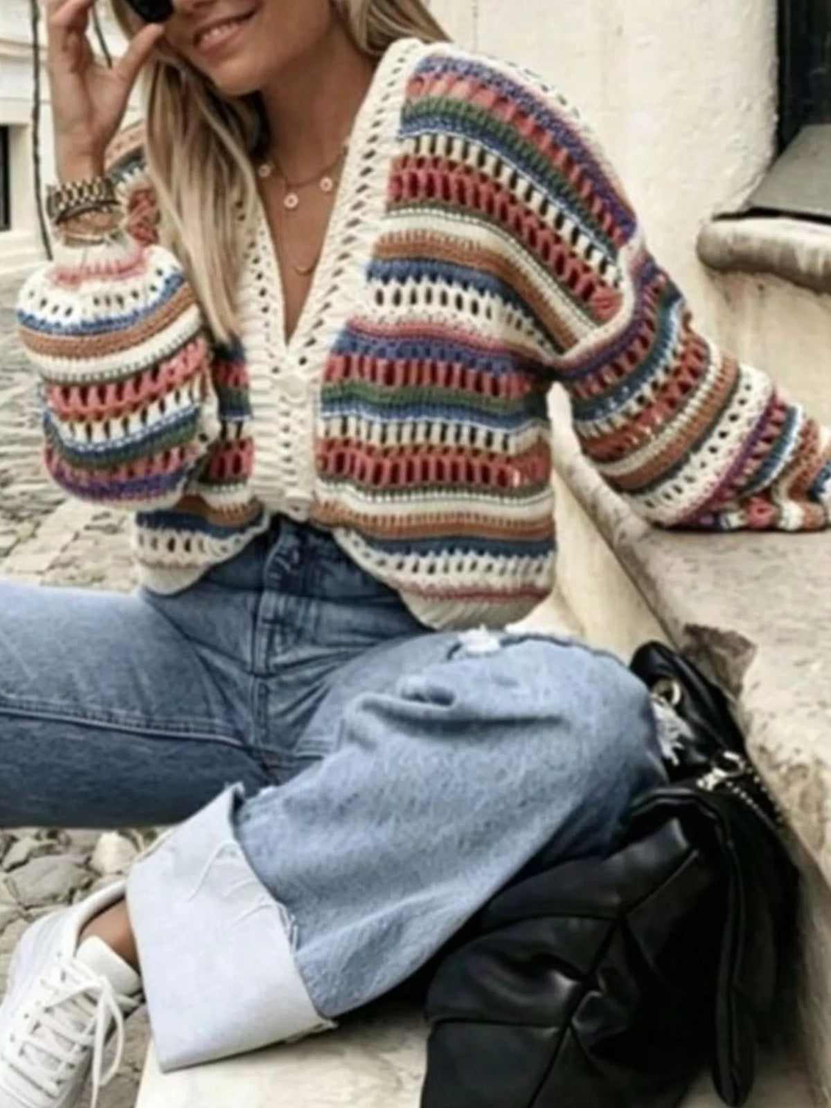 A woman sitting outdoors wearing a colorful striped crochet cardigan, light blue jeans, and white sneakers, with a black handbag beside her.