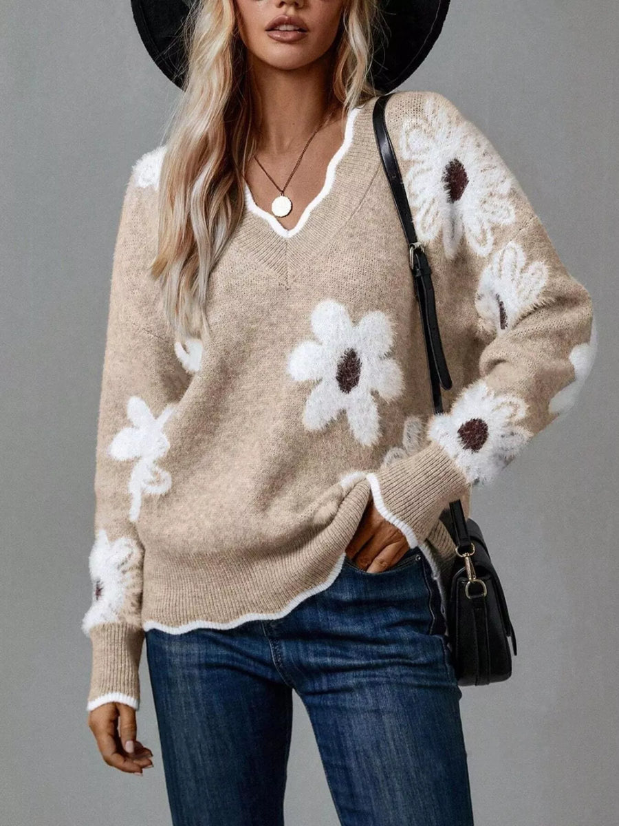 Woman wearing a beige sweater with white floral patterns, blue jeans, and a black hat against a gray background.