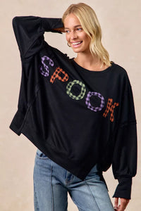 Woman wearing a black sweater with colorful 'SPOOK' text, standing against a beige background