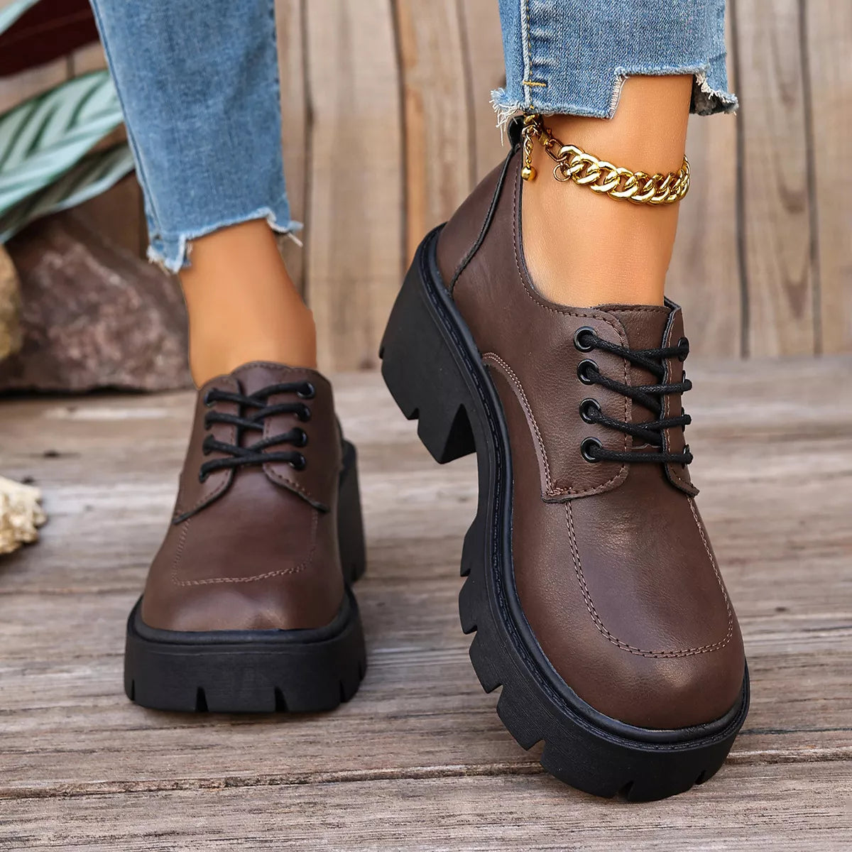 A pair of women's brown leather lace-up boots with chunky black platform soles, shown on a model wearing blue jeans and a gold chain anklet on a rustic wooden deck.