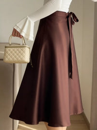 Person wearing a brown satin skirt holding a beige handbag indoors.