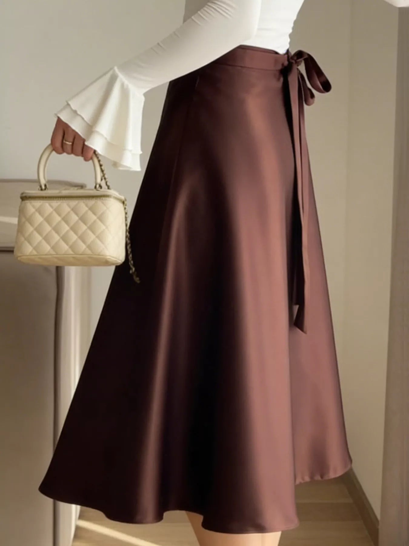 Person wearing a brown satin skirt holding a beige handbag indoors.