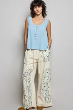 Woman wearing a light blue sleeveless top and cream pants with floral embroidery on a gray background