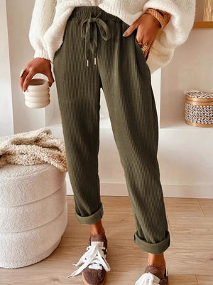 Person wearing green ribbed pants with a white sweater and brown shoes, holding a cup.