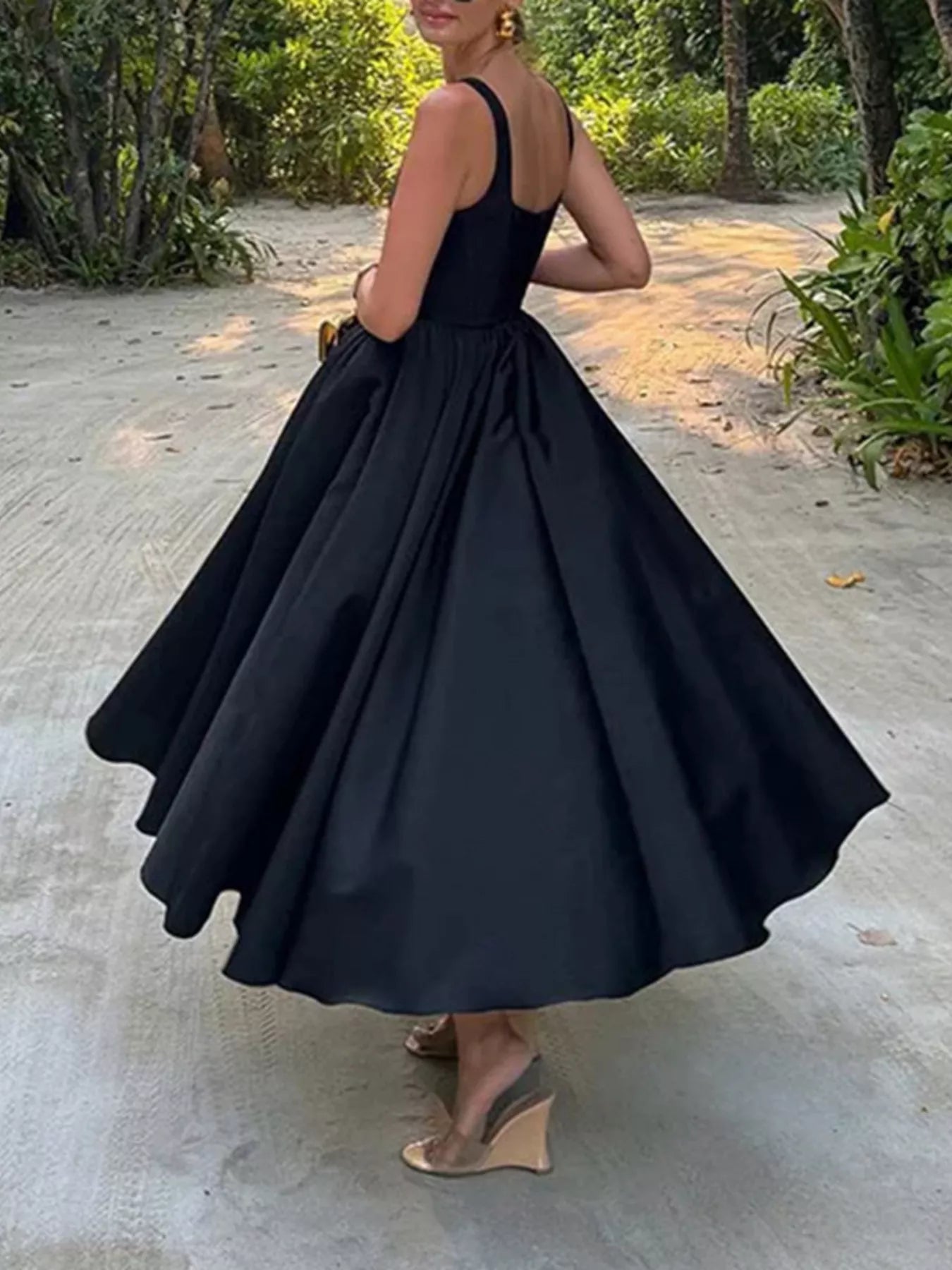 Woman wearing a black dress standing outdoors on a concrete surface with greenery in the background.
