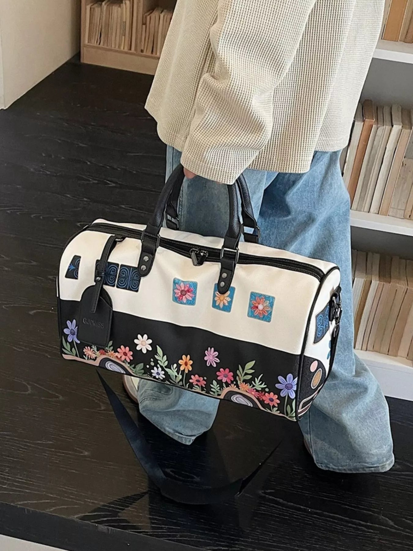 Person holding a floral-patterned duffel bag in a room with wooden flooring.