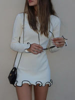 Woman wearing a white dress with black trim holding sunglasses against a plain background