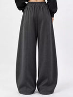 A person wearing loose, wide-leg dark gray sweatpants with an elastic waistband, paired with a black long-sleeve top, shown from the back against a plain light gray studio background.