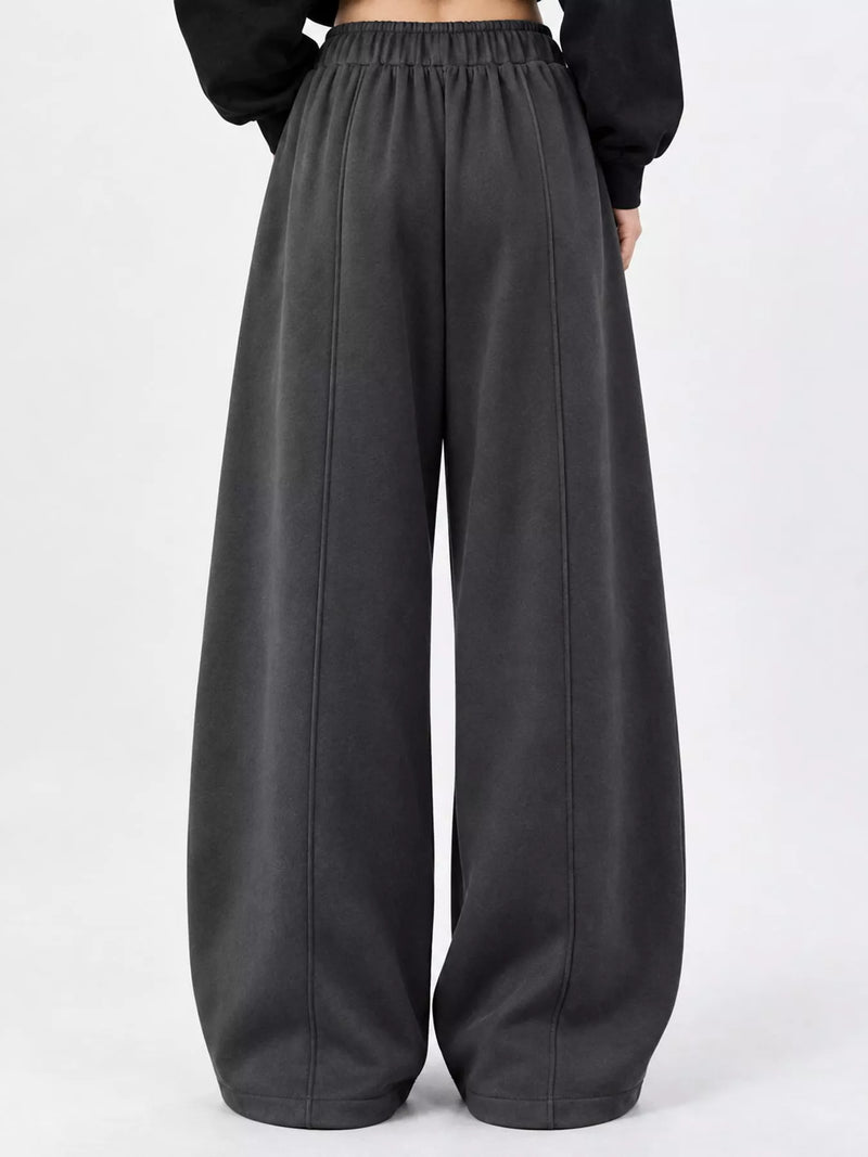 A person wearing loose, wide-leg dark gray sweatpants with an elastic waistband, paired with a black long-sleeve top, shown from the back against a plain light gray studio background.
