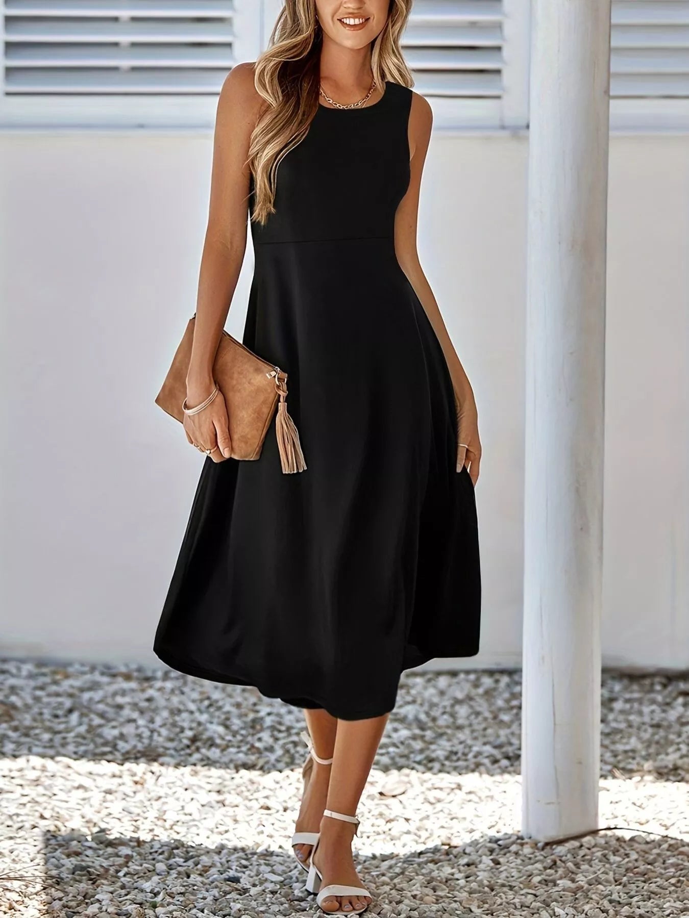 Woman wearing a black sleeveless dress holding a brown clutch against a white wall.