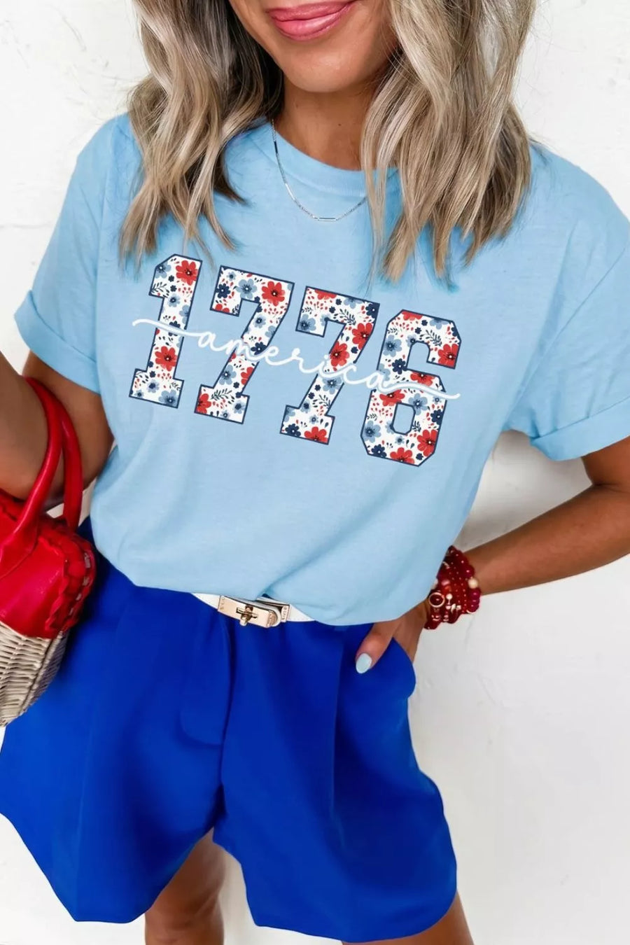 Woman wearing a light blue t-shirt with '1776' design, holding a red handbag.