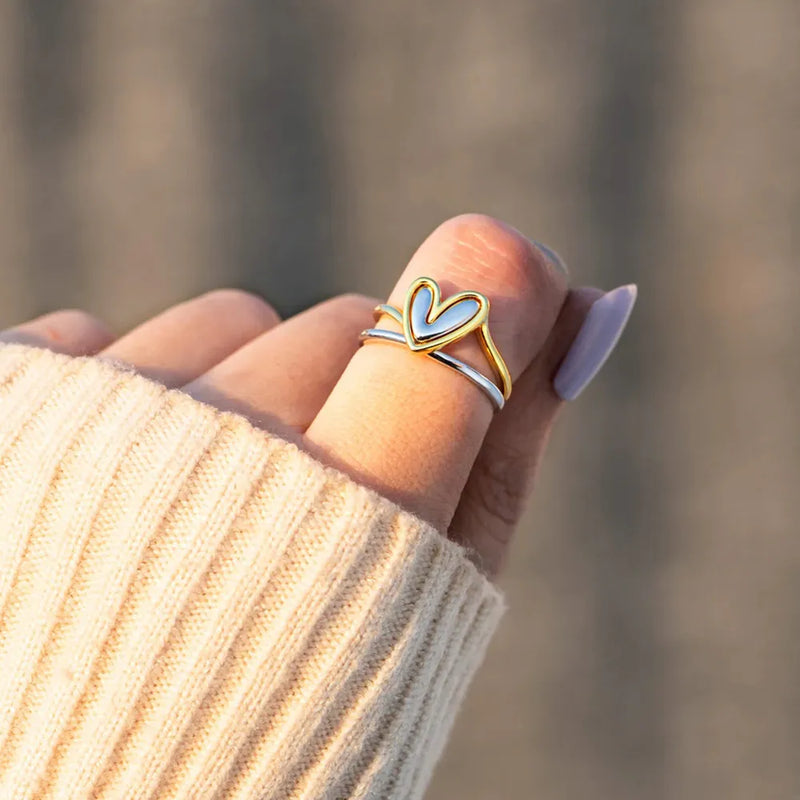 Hand wearing a gold and silver heart-shaped ring with blurred background