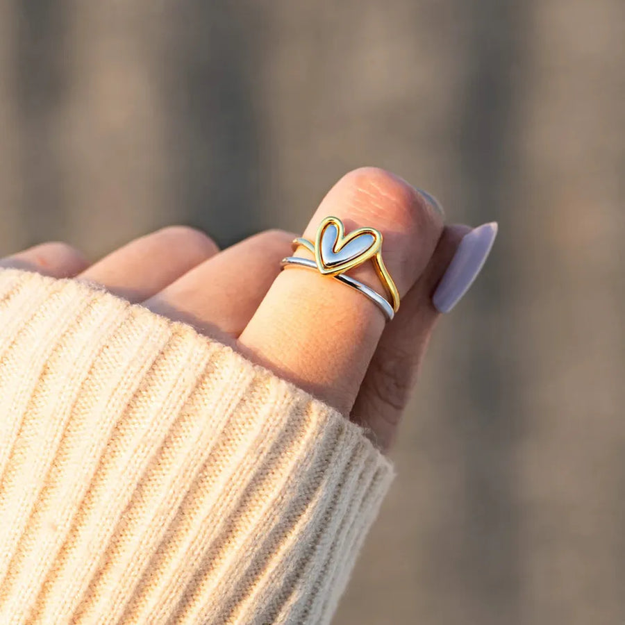 Hand wearing a gold and silver heart-shaped ring with blurred background
