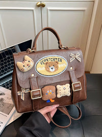 Brown handbag with bear-themed decorations held by a person, with a laptop and newspaper in the background.