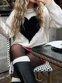 Person wearing a white sweater with a black heart design, sitting outdoors.