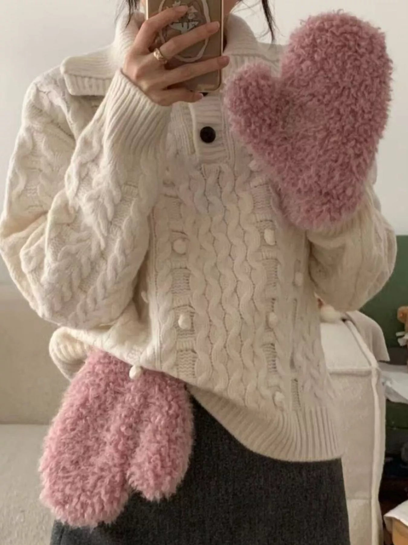 Person wearing a white cable knit sweater and pink fuzzy mittens indoors.