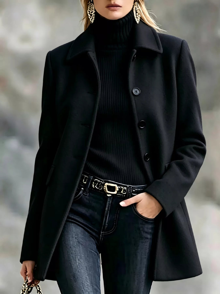 A woman wearing a black wool-blend coat over a black ribbed turtleneck sweater, paired with dark blue jeans and a gold-tone belt, photographed against a blurred gray outdoor background.