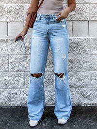 Person wearing light blue ripped jeans standing against a stone wall.