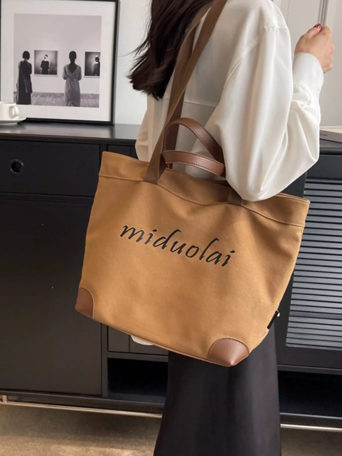 Person holding a tan tote bag with 'miduolai' branding in an indoor setting.