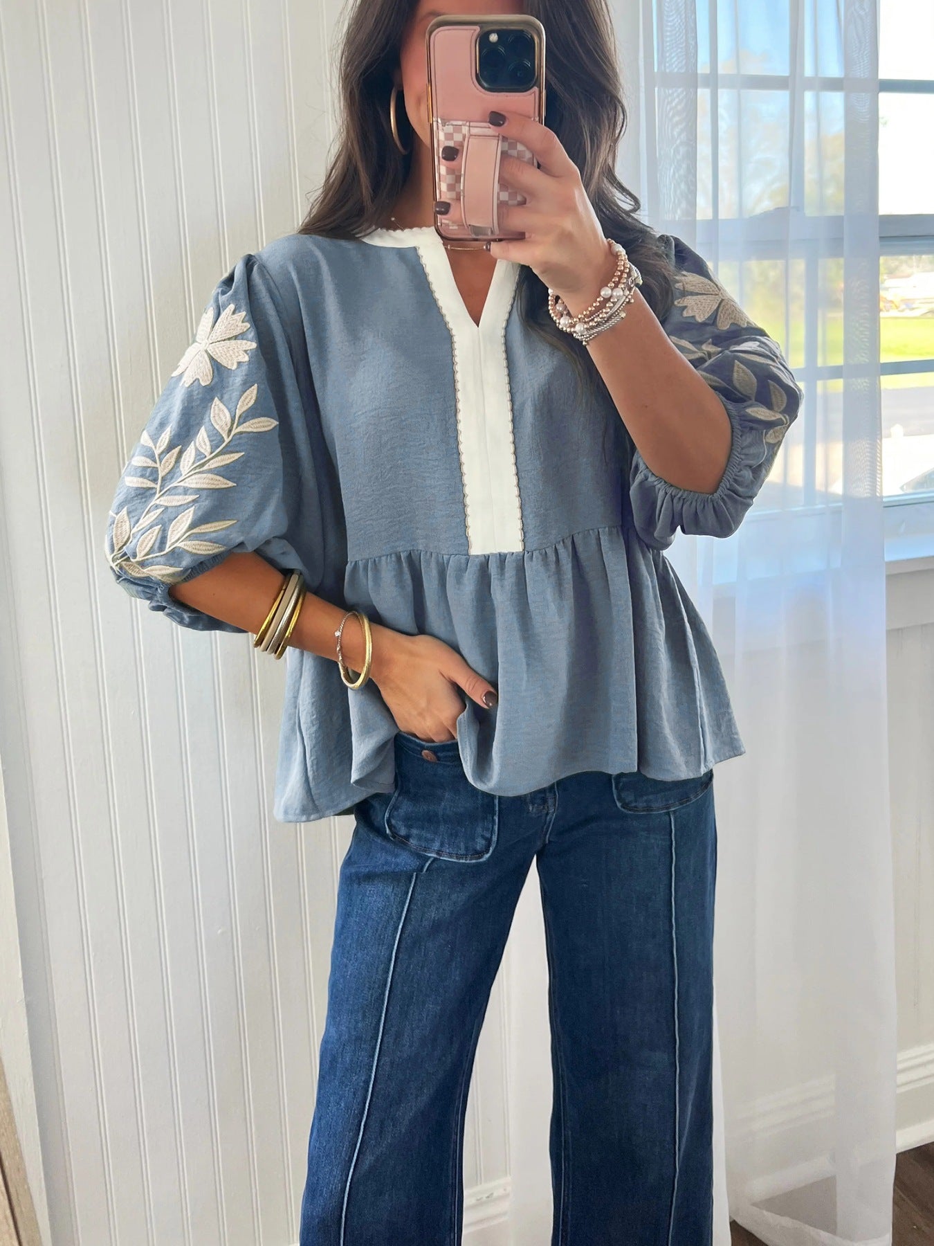 Person wearing a blue top with floral embroidery and jeans indoors.