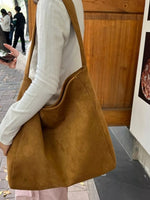 Person holding a brown leather bag outdoors with a wooden door and people in the background