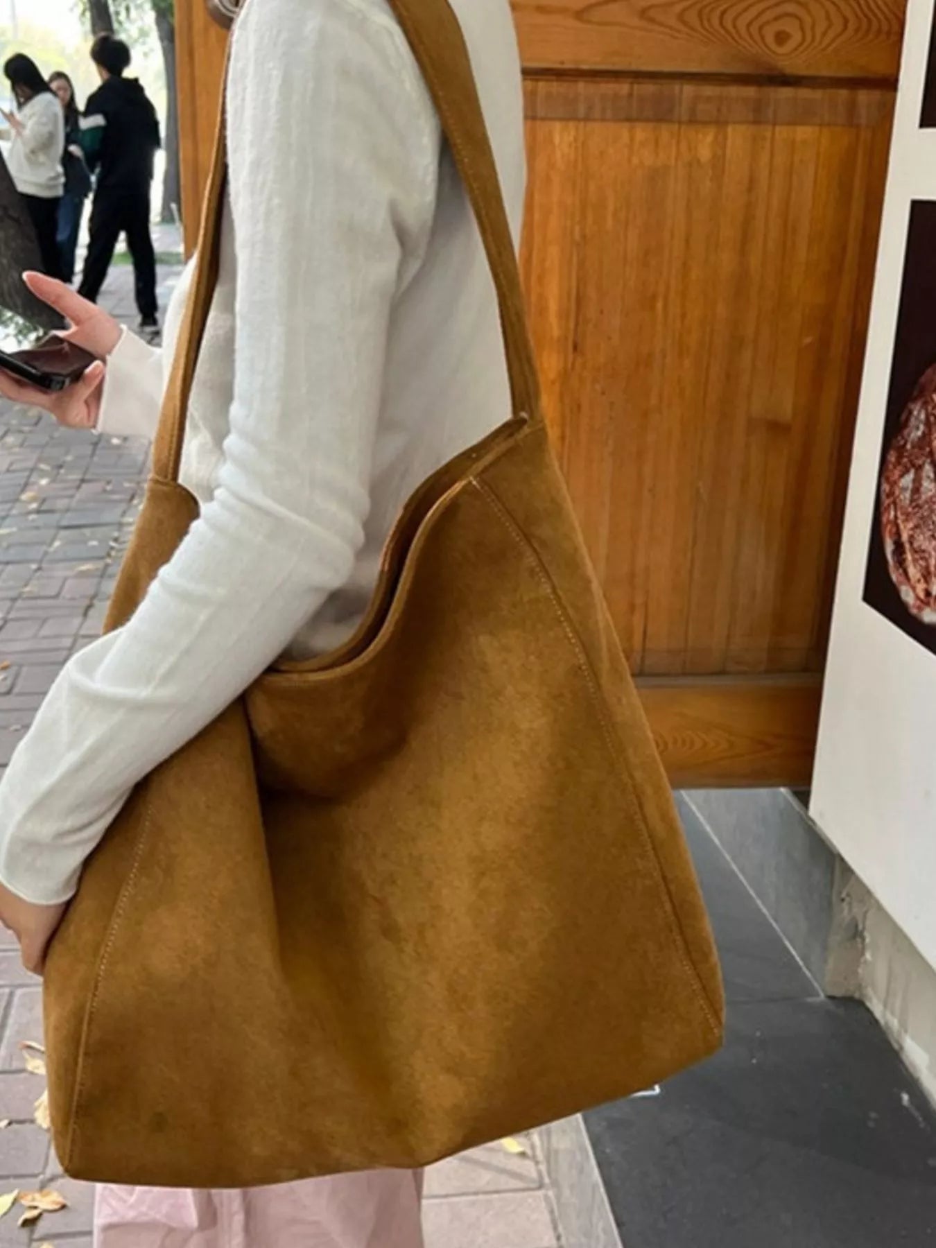 Person holding a brown leather bag outdoors with a wooden door and people in the background