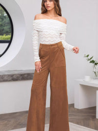 Woman wearing a white off-shoulder top and brown wide-leg pants in a modern indoor setting.