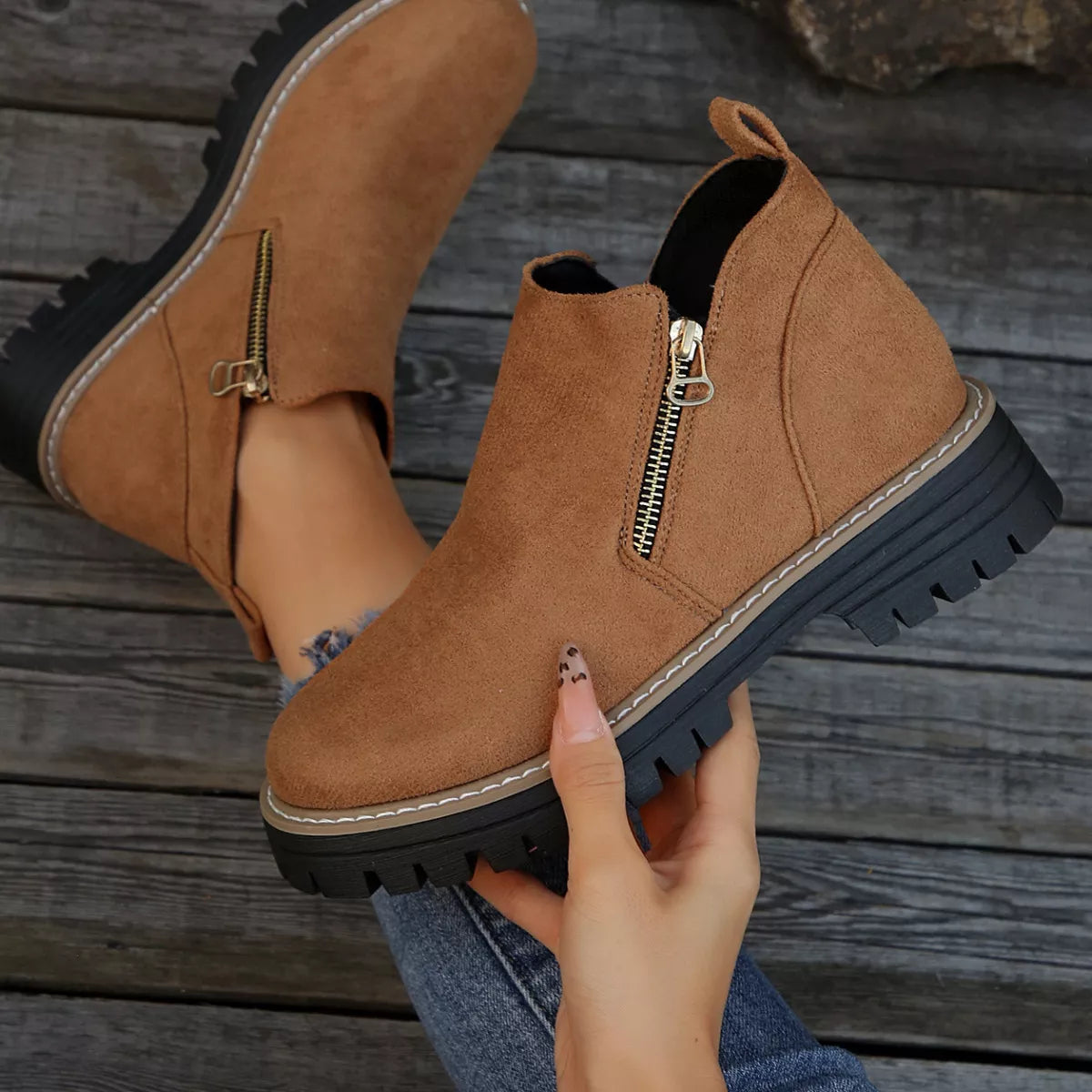 Brown suede ankle boots with zippers held by a hand on a wooden surface