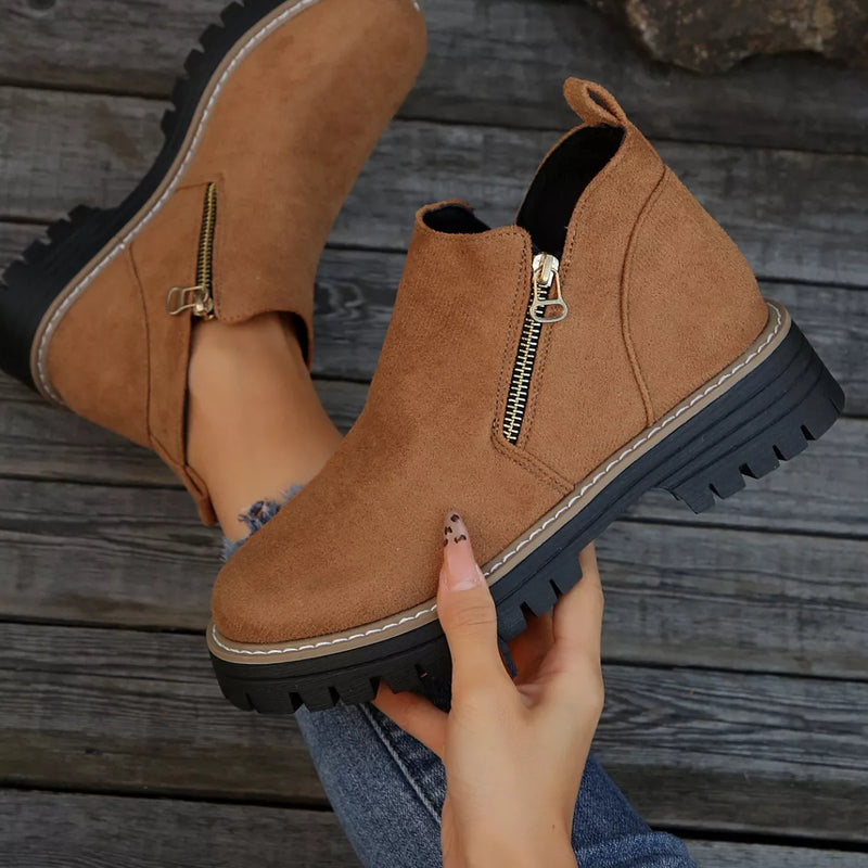 Brown suede ankle boots with zippers held by a hand on a wooden surface