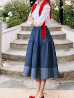 Person wearing a white shirt, red vest, and blue skirt standing on stone steps with greenery in the background.