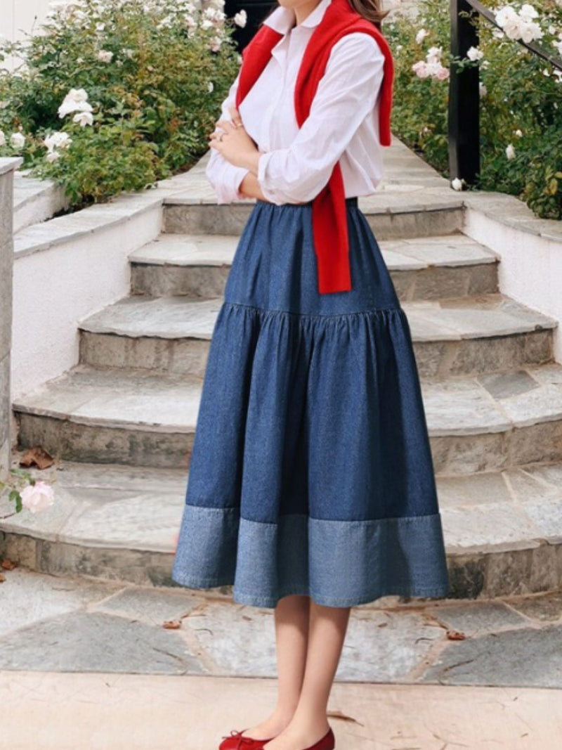 Person wearing a white shirt, red vest, and blue skirt standing on stone steps with greenery in the background.