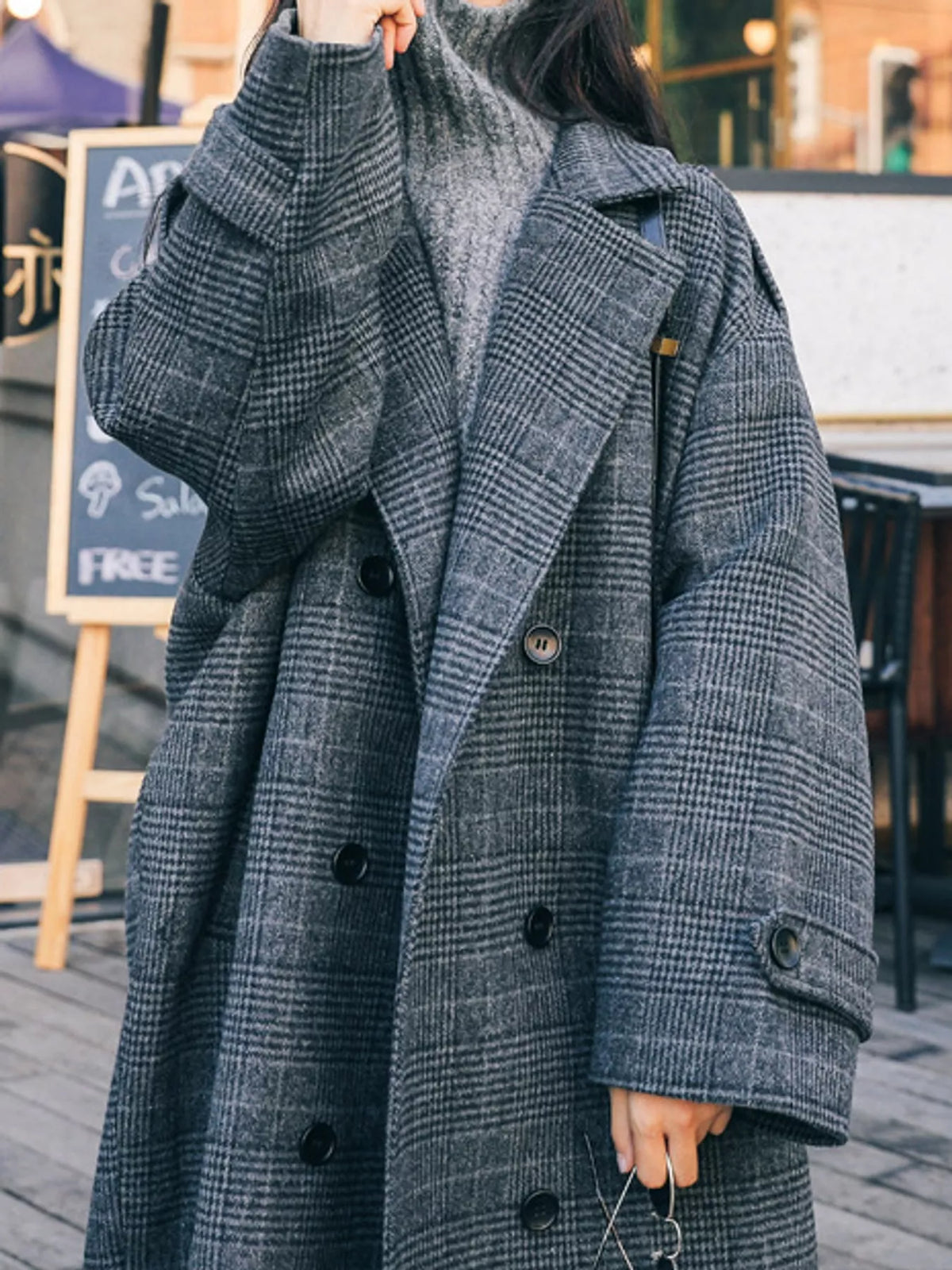 A person wearing an oversized double-breasted plaid coat in dark gray and black, layered over a gray knit turtleneck, photographed outdoors on a street or patio setting.