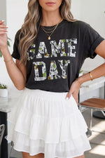 Woman wearing a black 'GAME DAY' t-shirt and white skirt indoors.