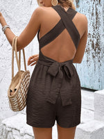 Woman wearing a brown open-back romper with a textured surface, holding a woven bag against a textured wall.