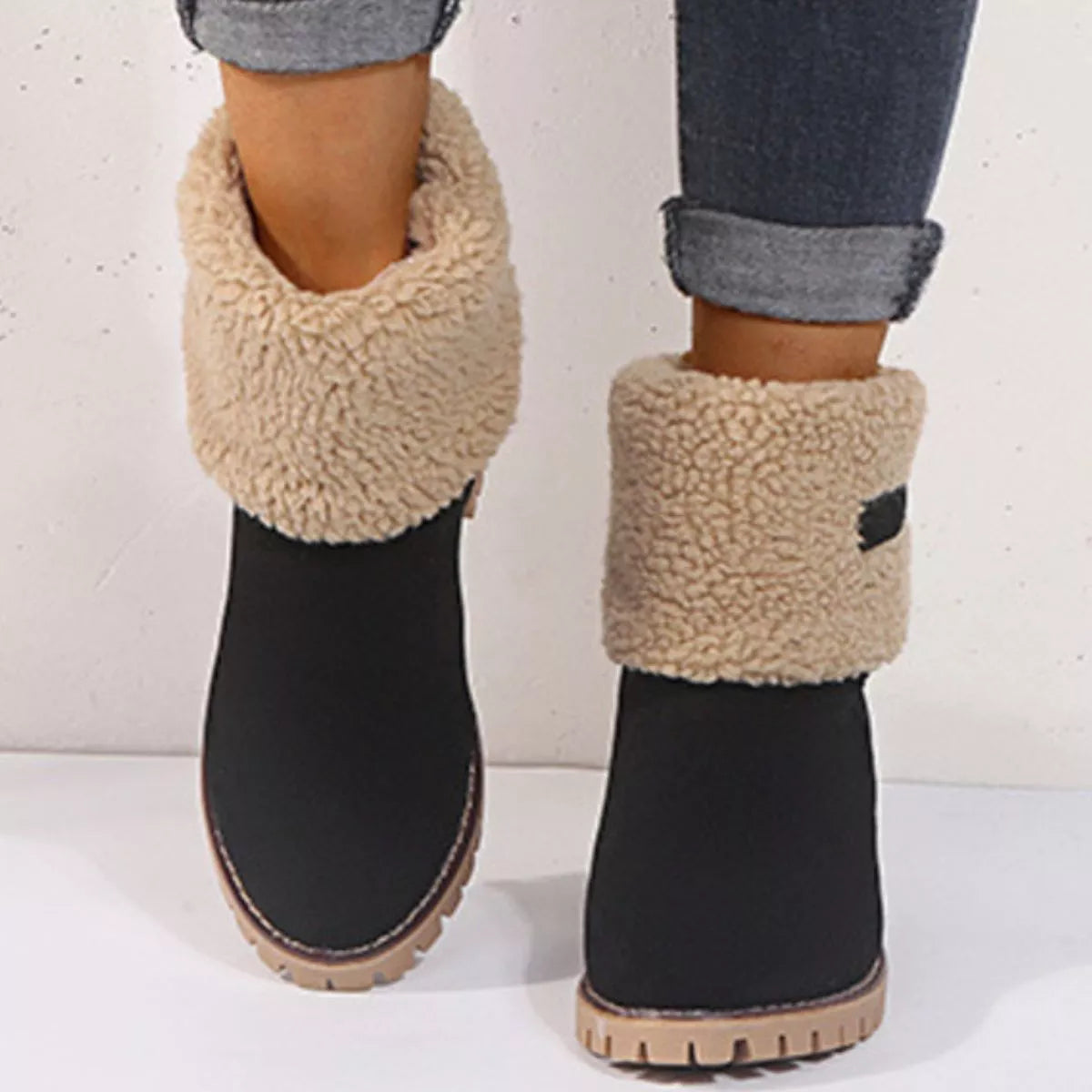 Black snow boots with beige fuzzy cuffs worn by a person on a white background