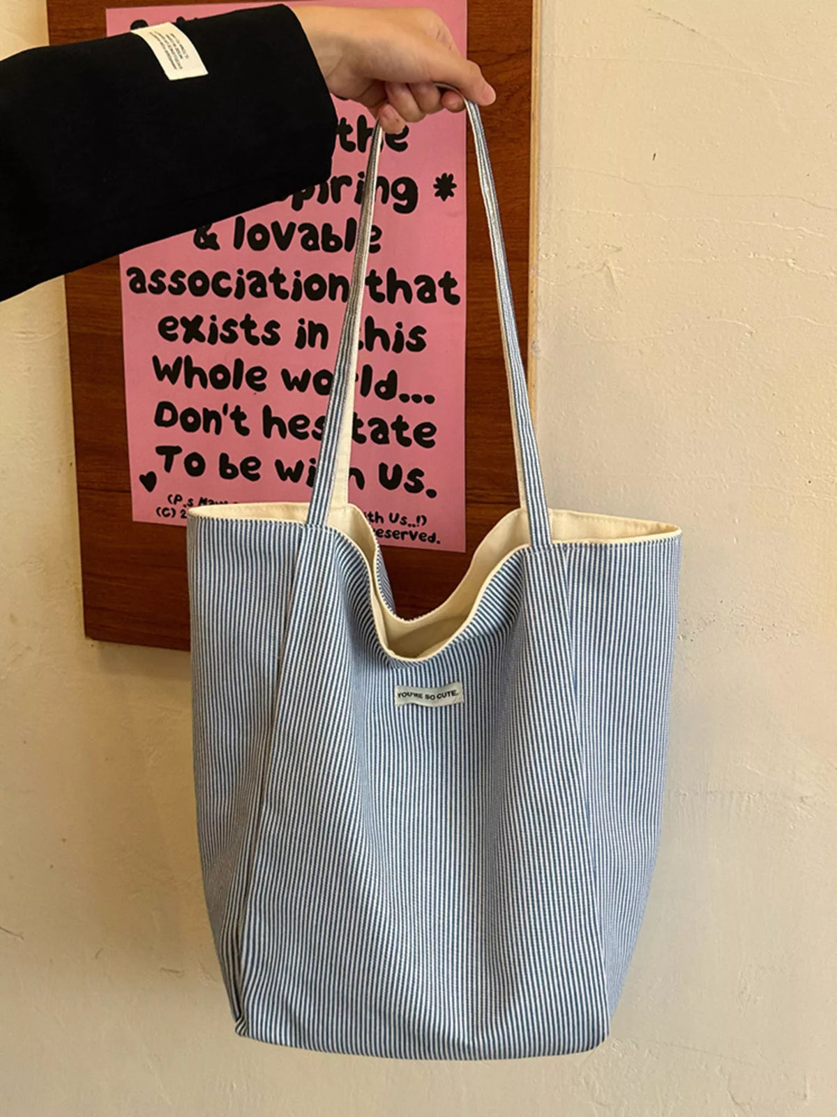 Blue striped tote bag held by a person against a wall with text.