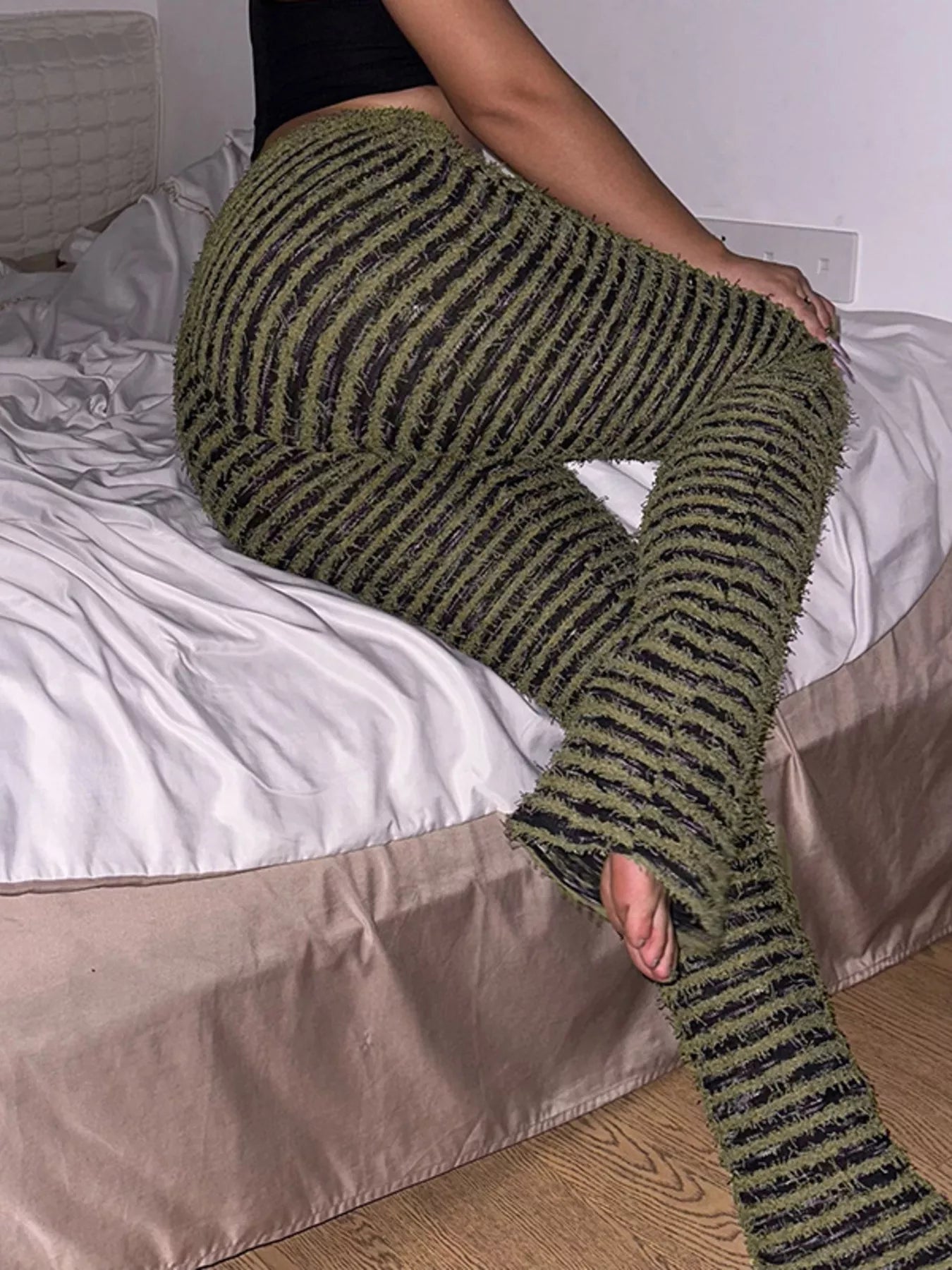 A person sitting on a bed wearing chunky olive green and black striped fuzzy knit pants with open toes.